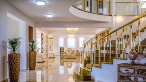 Ein großzügiger, gut beleuchteter Innenraum mit einem polierten Boden, einer imposanten Treppe mit goldfarbenen Geländern und einer modernen Wohnbereits mit Sofas und einem Chandelier.