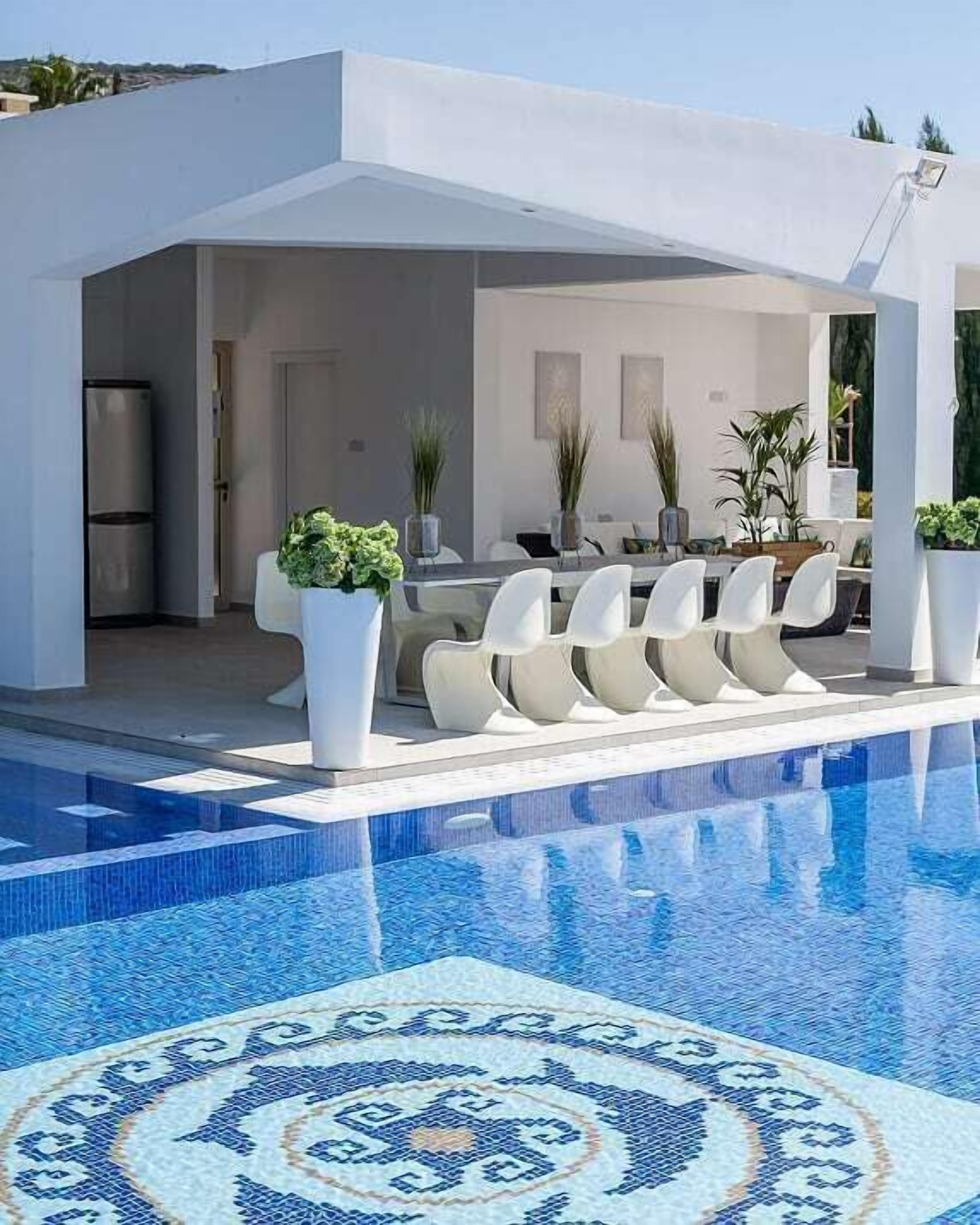 A modern outdoor pool area with a geometric tile pattern, a dining set with white chairs, and potted plants.