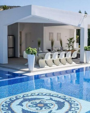 Un espace piscine extérieur moderne, agrémenté d’un motif géométrique en carreaux, d’un ensemble de mobilier de jardin avec des chaises blanches et de plantes en pot.