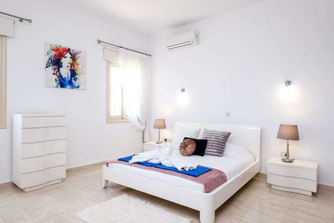 A modern bedroom with a white bed, bedside tables, a white dresser, a colorful abstract painting, and an air conditioning unit.