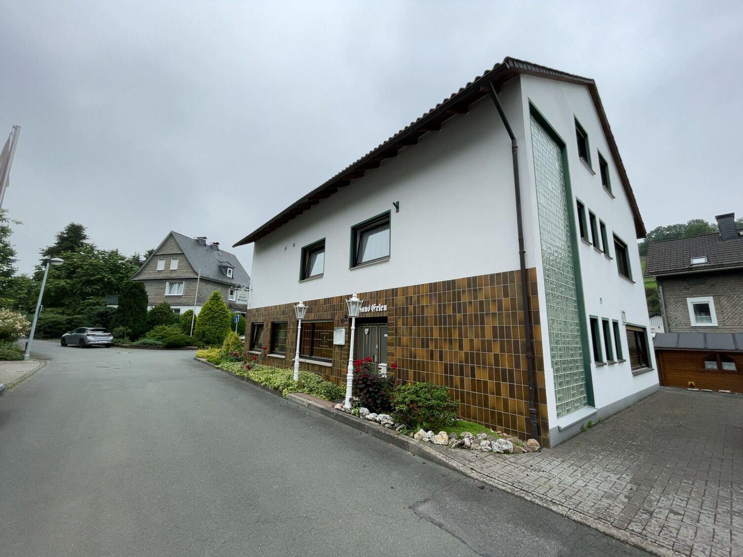 Um edifício de dois andares com uma placa 'Musterwaren' no andar térreo, um caminho de paralelepípedos e uma área residencial ao fundo.