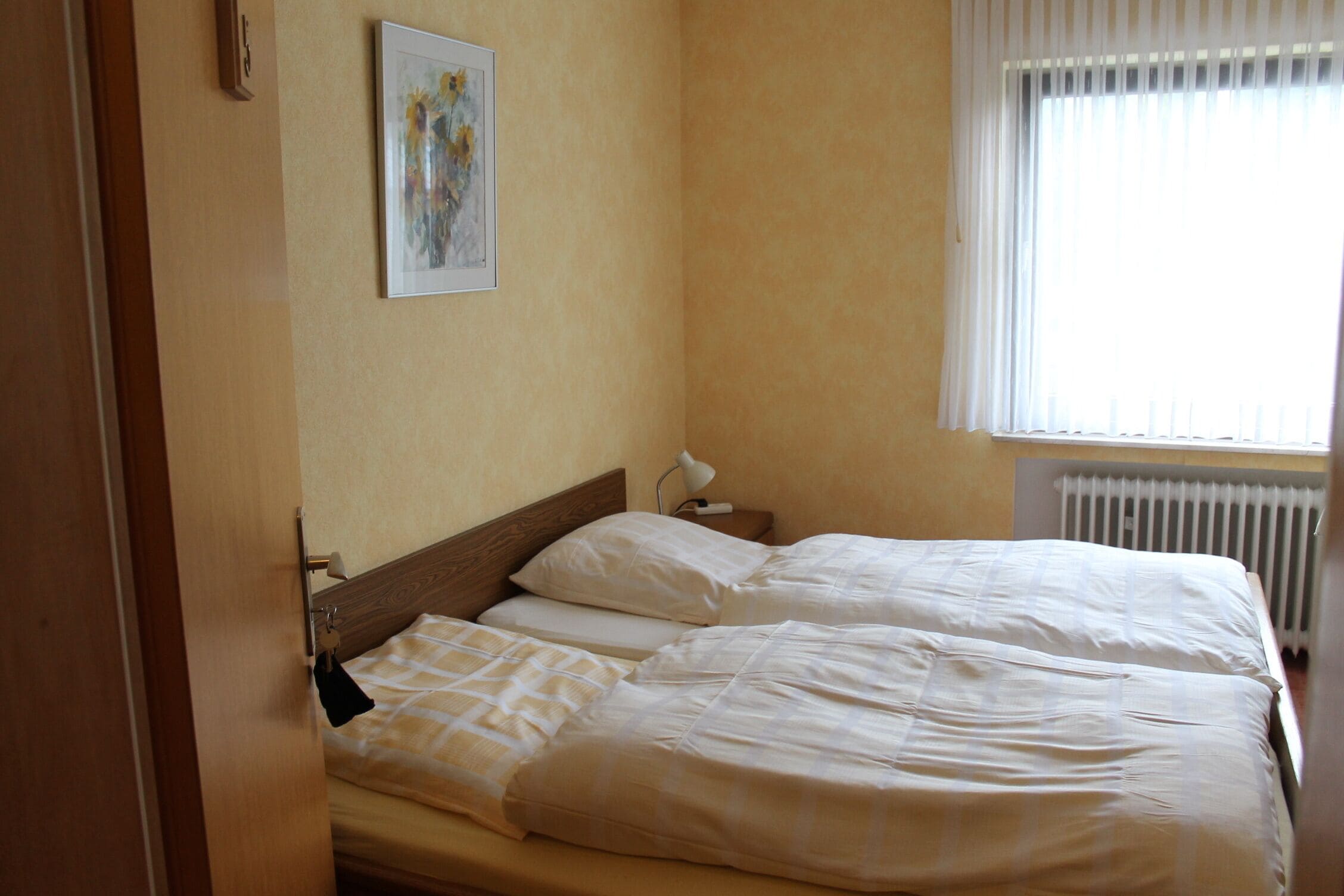 Habitación de hotel con cama, mesita de noche, lámpara, un cuadro en la pared y ventana con persianas.