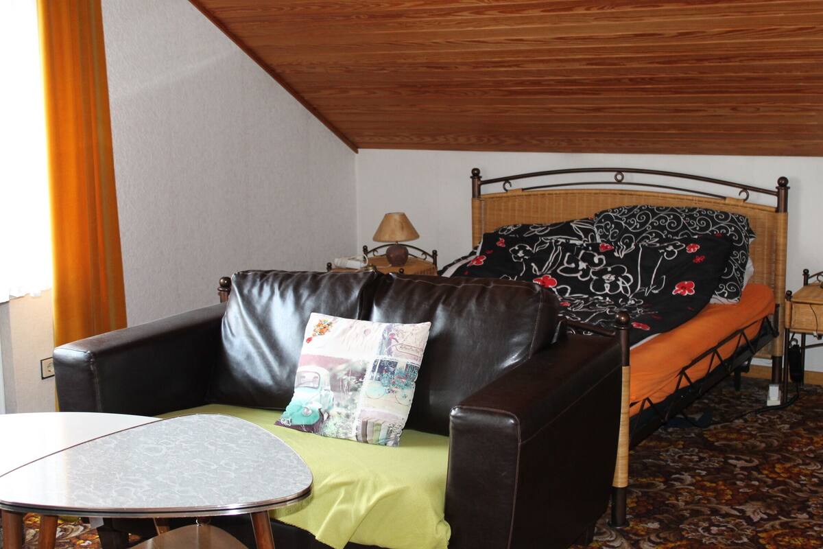 A room with a bed, a leather sofa, a small table, and a lamp.