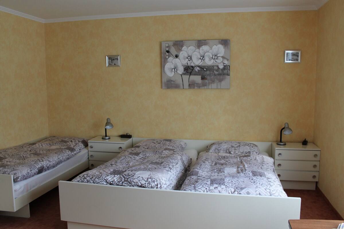 A bedroom with two single beds, a larger bed, a painting on the wall, and bedside tables with lamps.