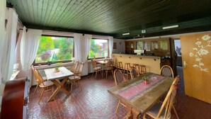 A dining area with wooden furniture, a kitchen in the background, and large windows. - Hotel Haus Erlen (Olsberg)