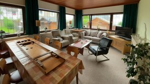 A spacious living room with a wooden ceiling, a sofa set, a dining table with a board game, a TV, and large windows with curtains. - Hotel Haus Erlen (Olsberg)
