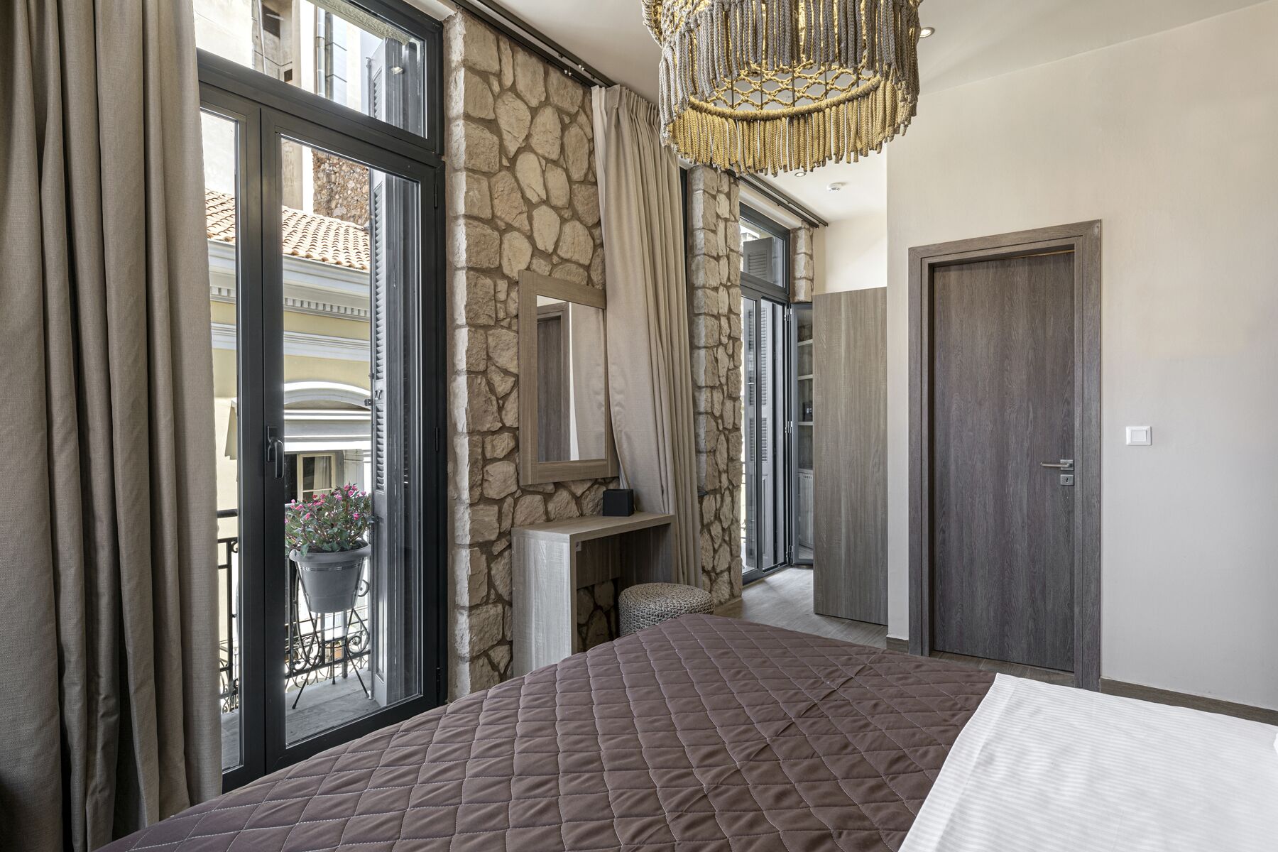 A bedroom with a large bed, stone walls, a chandelier, and a view of the outside balcony.