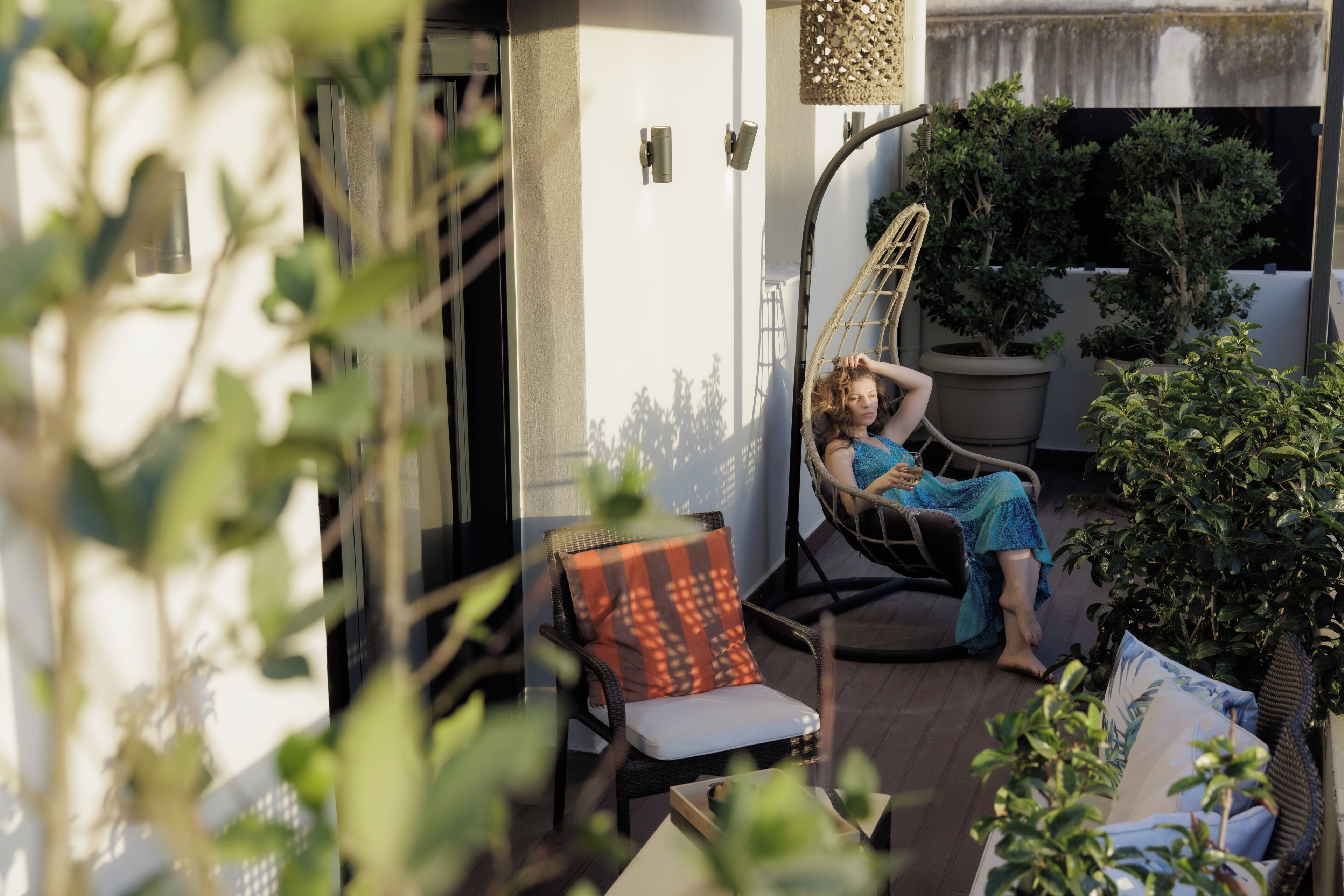 A person relaxing on a wicker chair with a cushion, a potted plant, and a hanging light fixture in a sunlit outdoor area.