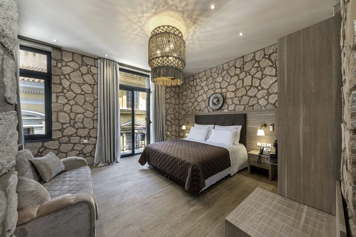 Une chambre avec un mur en pierre, un grand lit, un lustre, une table de chevet avec un téléphone et une vue sur le balcon extérieur.