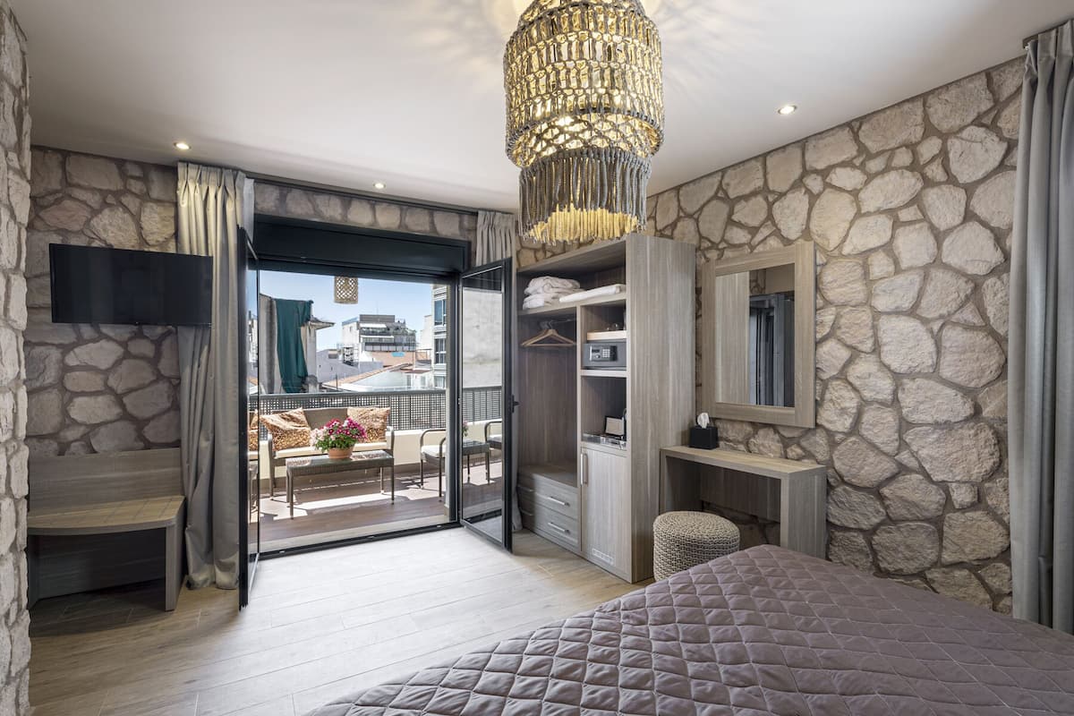 Une chambre avec un mur en pierre, un lit, une armoire, un bureau et un balcon donnant sur des immeubles.