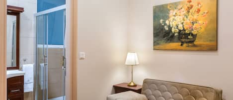 A hotel room with a glass shower, a beige sofa, a bedside table with a lamp, and a painting of flowers on the wall.