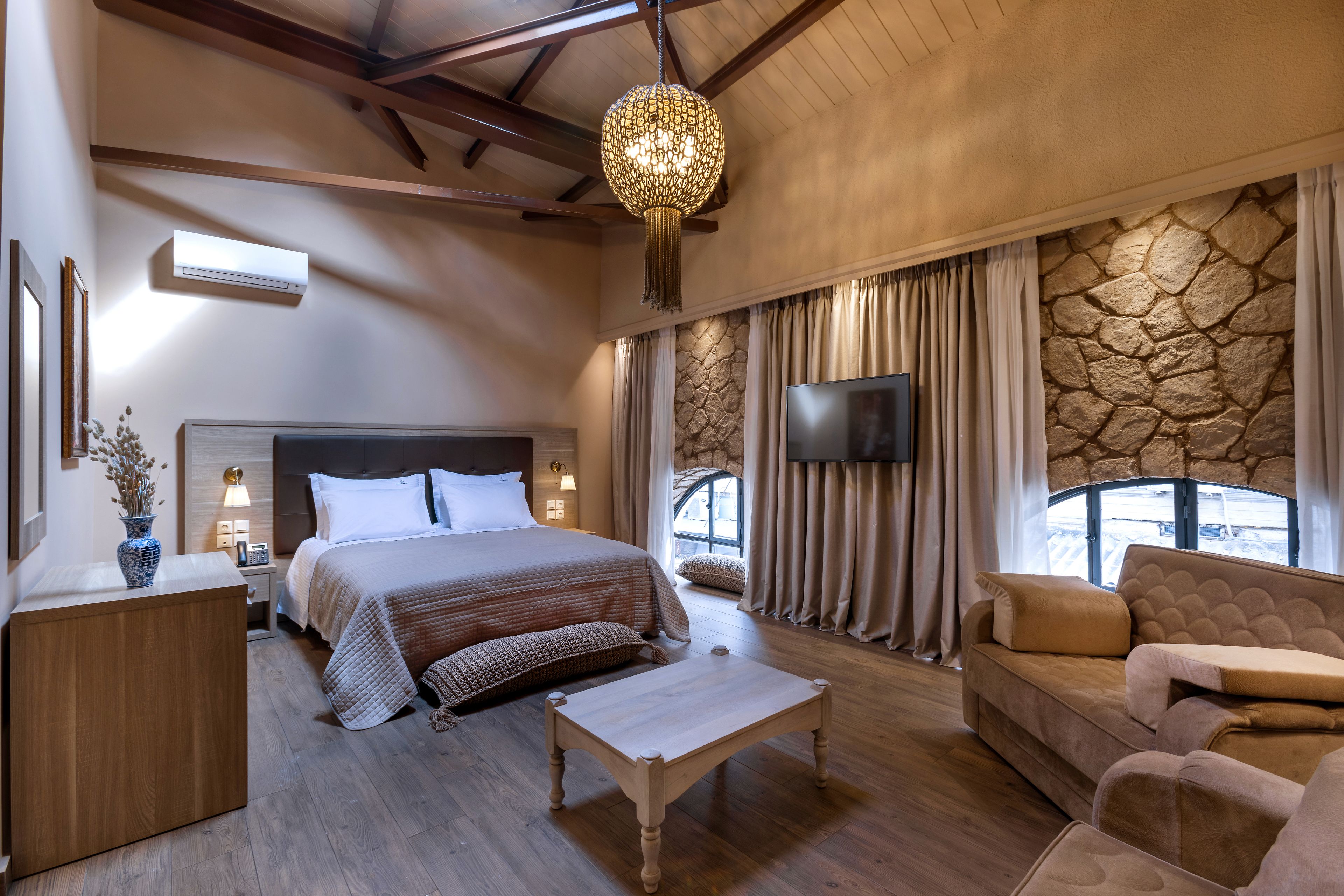 A spacious bedroom with a large bed, a wooden nightstand, a bedside table, a flat-screen TV, a stone accent wall, and a stone fireplace.