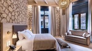 A hotel room with a stone wall, a bed with a headboard, a bedside table with a phone, a sofa, and a large window with curtains. - Athens Manor Houses Suites Apartments (Athens)