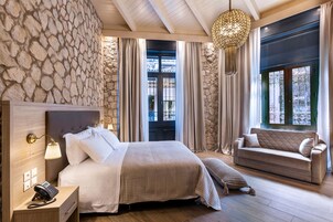 A hotel room with a stone wall, a bed with a headboard, a bedside table with a phone, a sofa, and a large window with curtains.