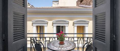 An open balcony with a round table and chairs, a potted plant, and a view of a building with arched windows and yellow shutters.