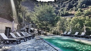 A pool area with lounge chairs and umbrellas, surrounded by trees and a mountainous backdrop. - L'Appollu (La Porta)