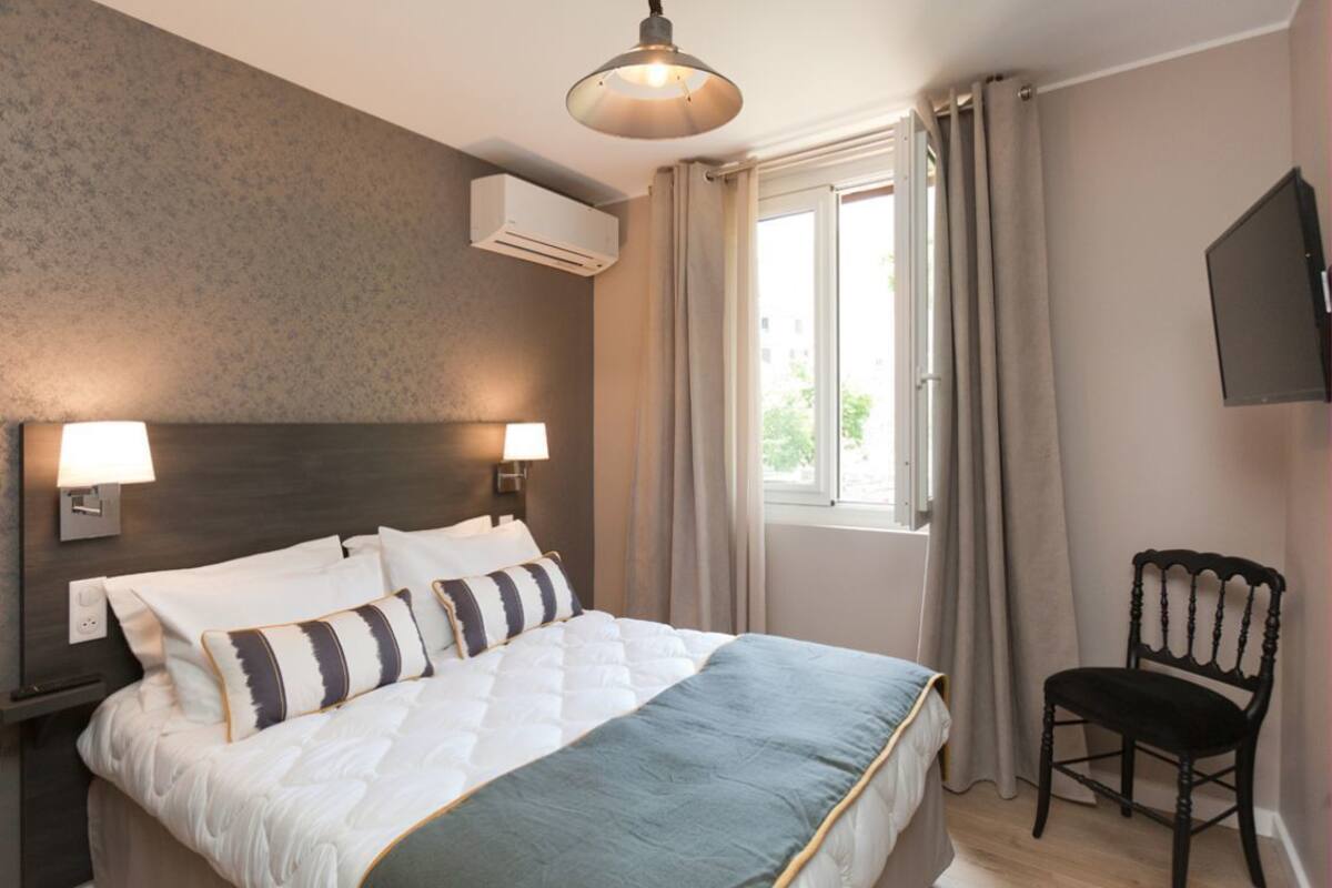 A hotel room with a large bed, two bedside lamps, a TV mounted on the wall, and a window with curtains.