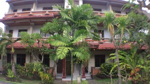 A multi-story building with a red-tiled roof, surrounded by lush greenery including palm trees and various shrubs. - Mangga Bali Inn (Kuta)