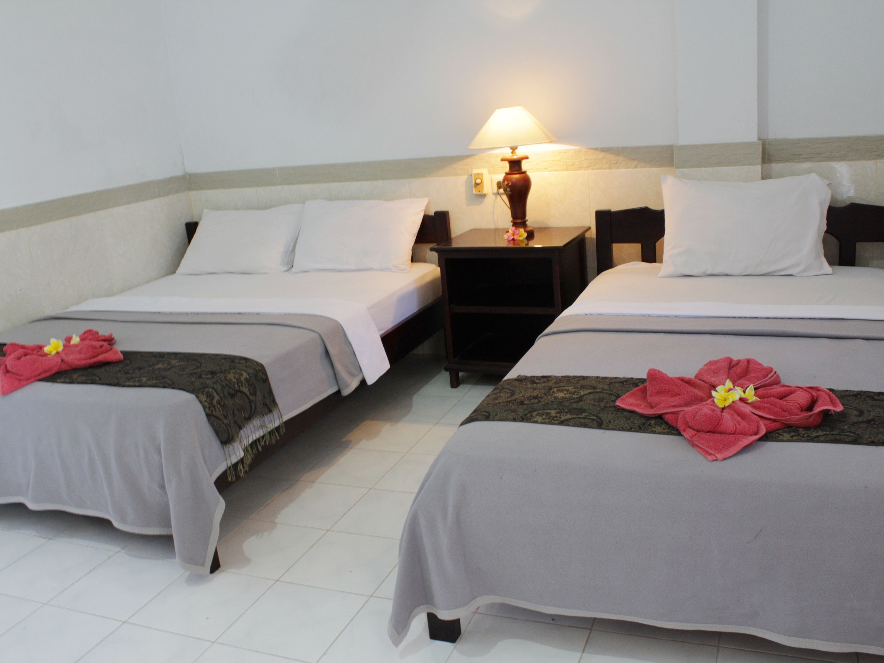 A hotel room with two beds, each with white linens and a red floral bedspread. There is a nightstand with a lamp between the beds.