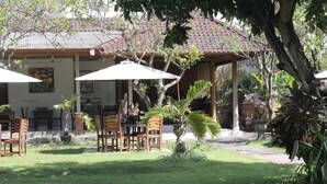 An outdoor seating area with wooden chairs and tables, surrounded by greenery and a building with a tiled roof. - Mangga Bali Inn (Kuta)