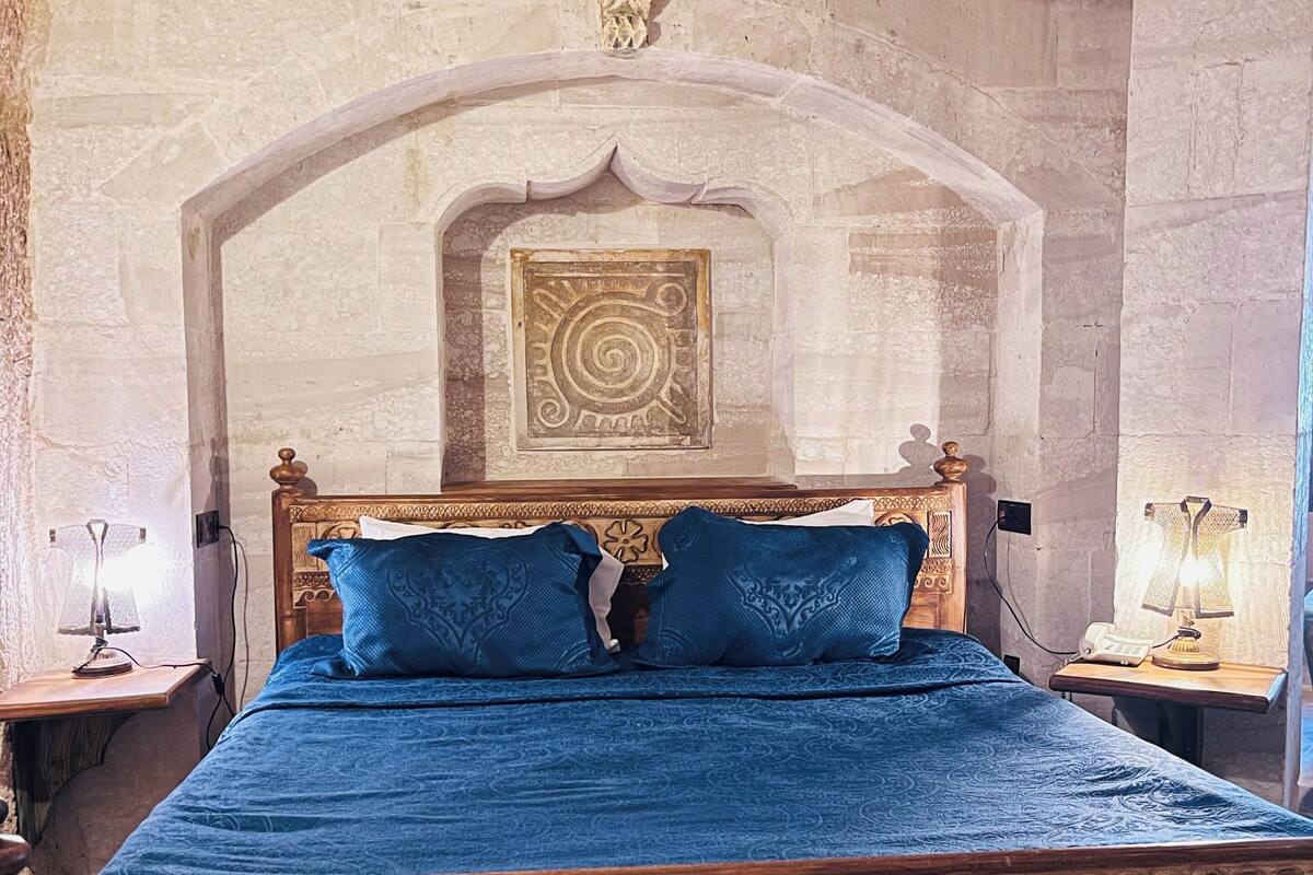 Una cama con colcha azul y dos almohadas, cabecera de madera con detalles florales, panel decorativo en la pared y techo con cortinas blancas.