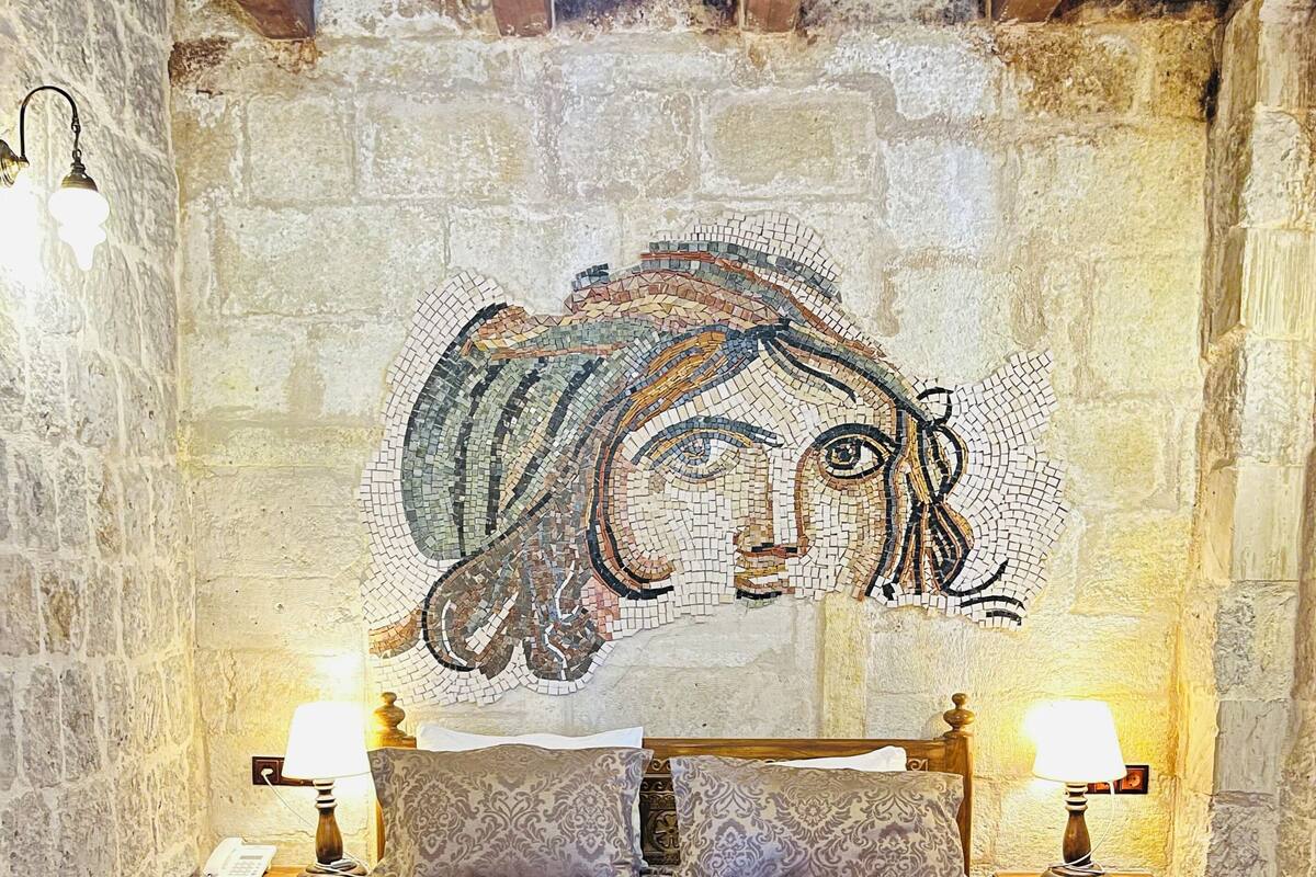 Un dormitorio con pared de piedra, una cama de madera con detalles florales, dos lámparas de noche y una obra de arte en mosaico sobre la cama.
