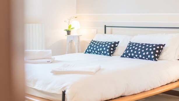 Un lit bien fait, avec une literie blanche et deux oreillers bleus à pois blancs, une table de chevet avec une lampe et un radiateur blanc.