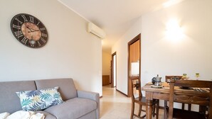 Un salon avec un canapé gris, un plaid à motifs, une table à manger en bois avec des chaises, et une horloge murale avec des chiffres romains.