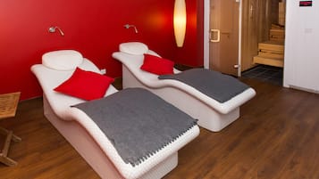 Una habitación con dos sillones de masaje, una pared roja, suelo de madera y la puerta de un sauna.