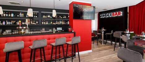 Un área de bar moderna con una pared de acento roja, varios taburetes y una variedad de botellas en estantes, además de un televisor de pantalla plana montado sobre la barra.
