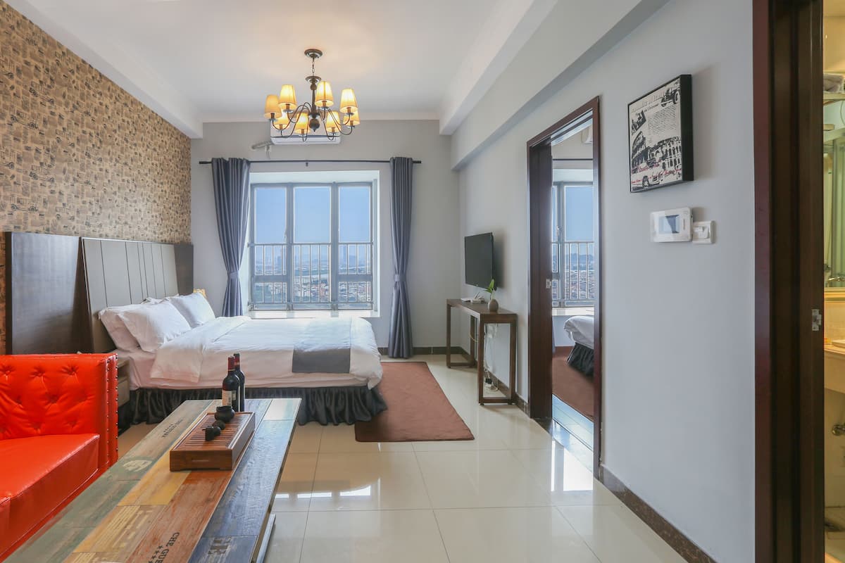 A hotel room with a bed, a red chair, a wooden table with a bottle and glasses, a TV, and a window with curtains.