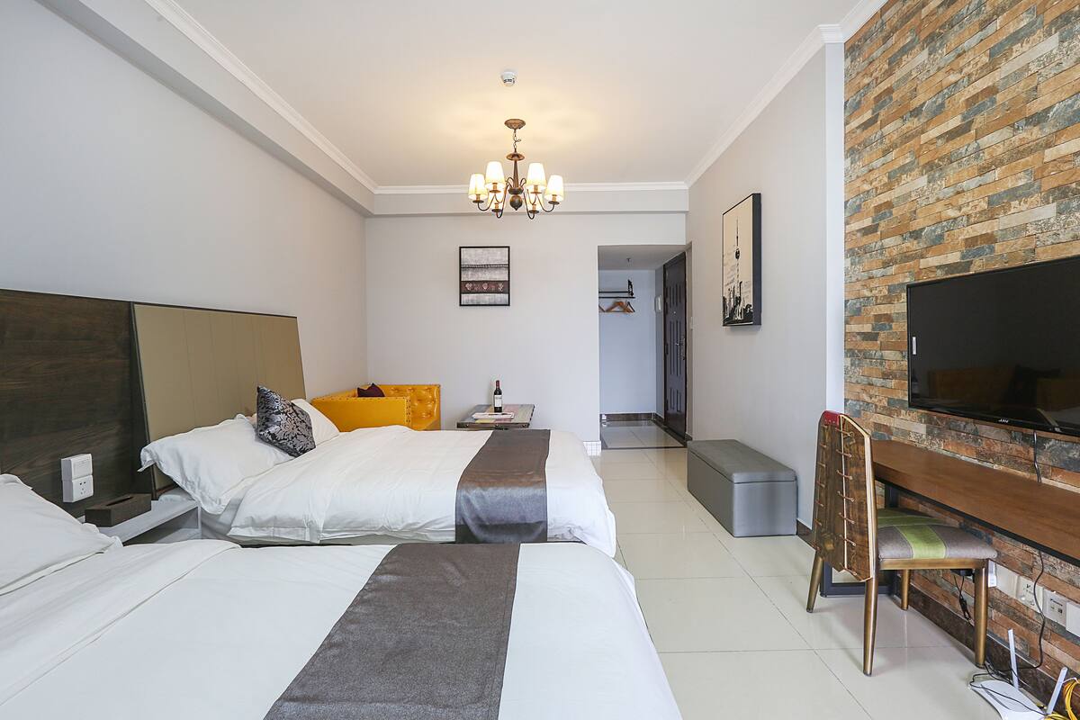 A hotel room with two beds, a TV, a desk, and a brick wall.