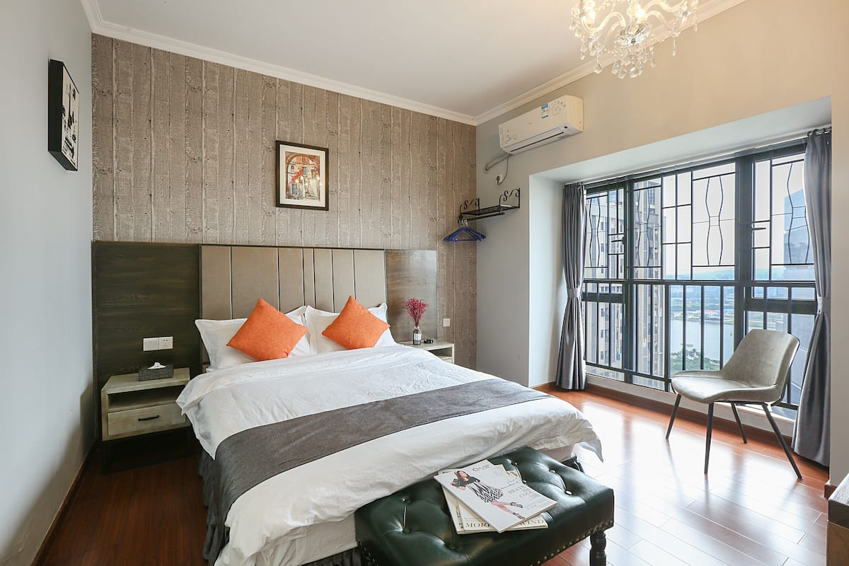 A bedroom with a large bed, a green ottoman, a nightstand, a wall-mounted air conditioner, and a view of a cityscape through a window.