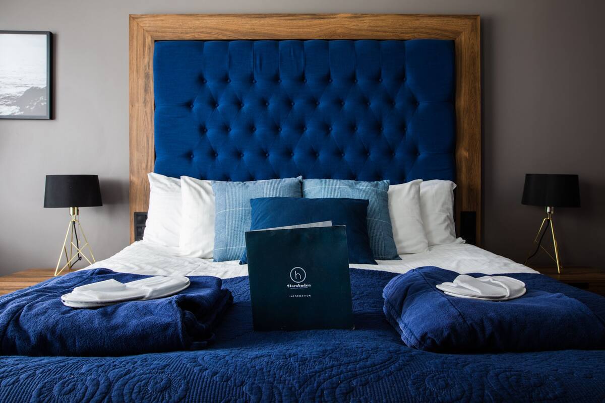 Ein ordentlich gemachtes Bett mit blauem Kopfteil, weißen Kissen und einem gefalteten Handtuch. Auf dem Bett liegt ein dunkel gefärbtes Buch oder eine Ordner.