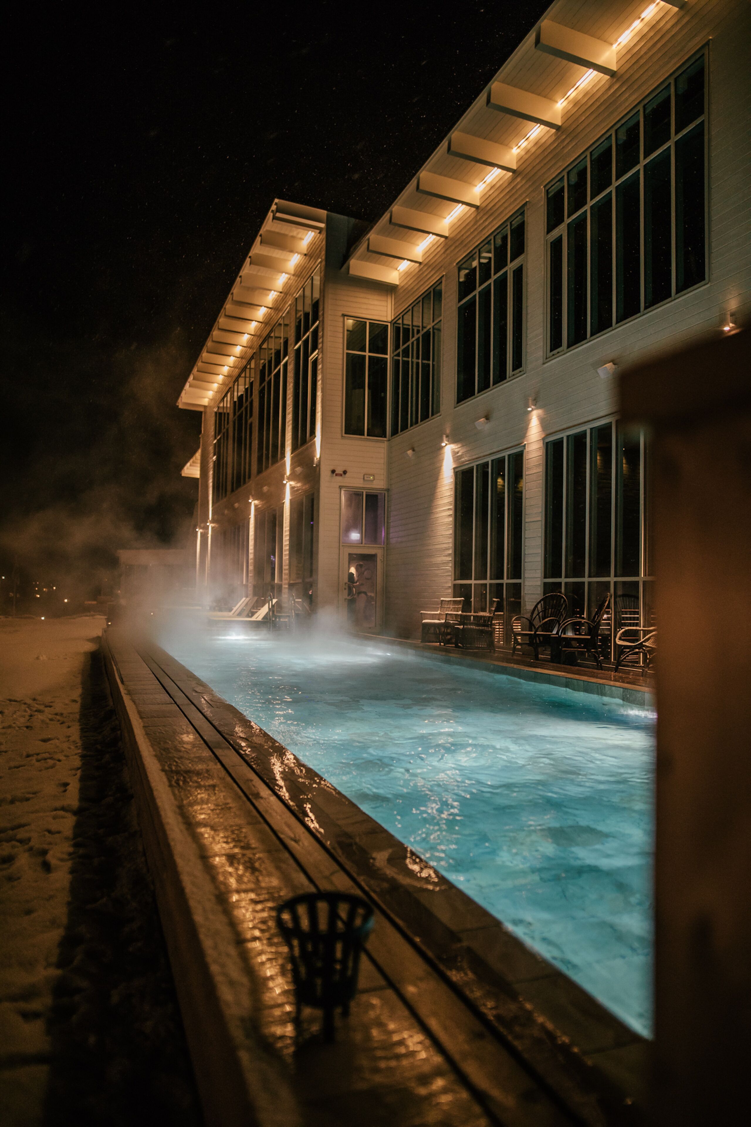 A nighttime view of a hotel with a heated outdoor swimming pool, a covered deck area, and large windows.