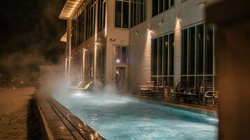 A nighttime view of a hotel with a heated outdoor swimming pool, a covered deck area, and large windows.