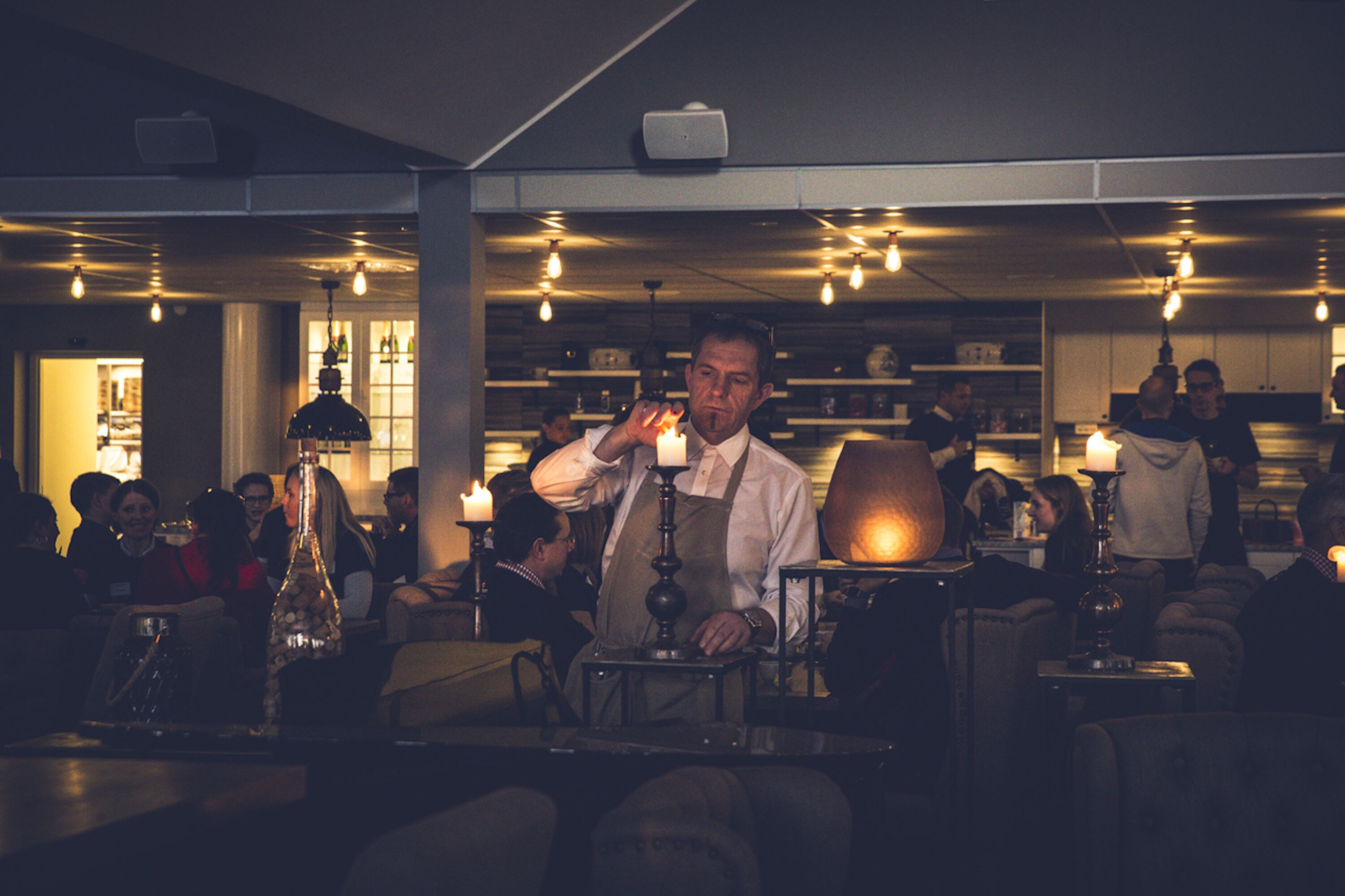 En svagt upplyst bar med gäster sittande vid borden, en bartender som blandar drinkar och en varm och inbjudande atmosfär med hängande lampor.