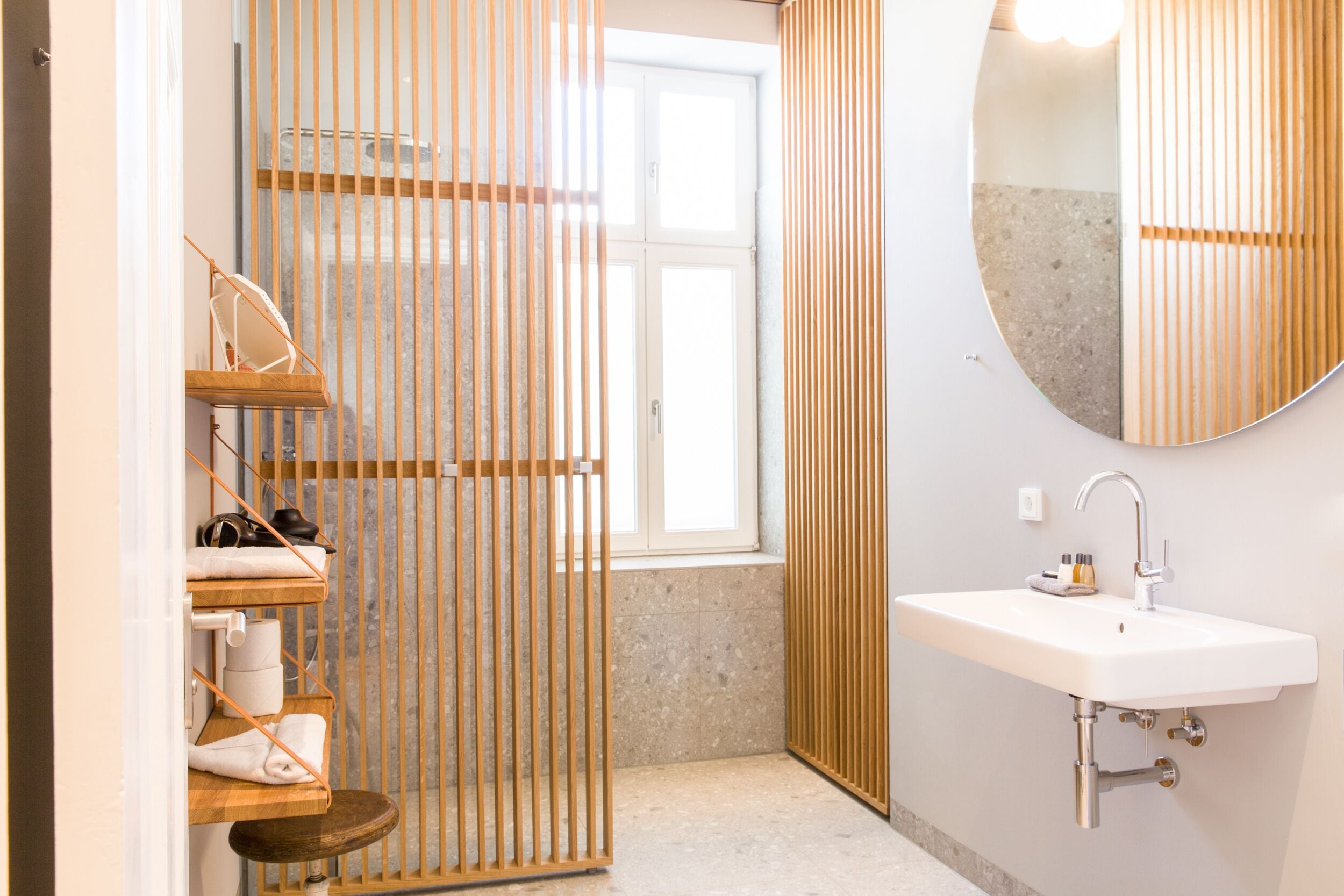 A modern bathroom with a wooden partition, a round mirror, a white sink, and a small stool.