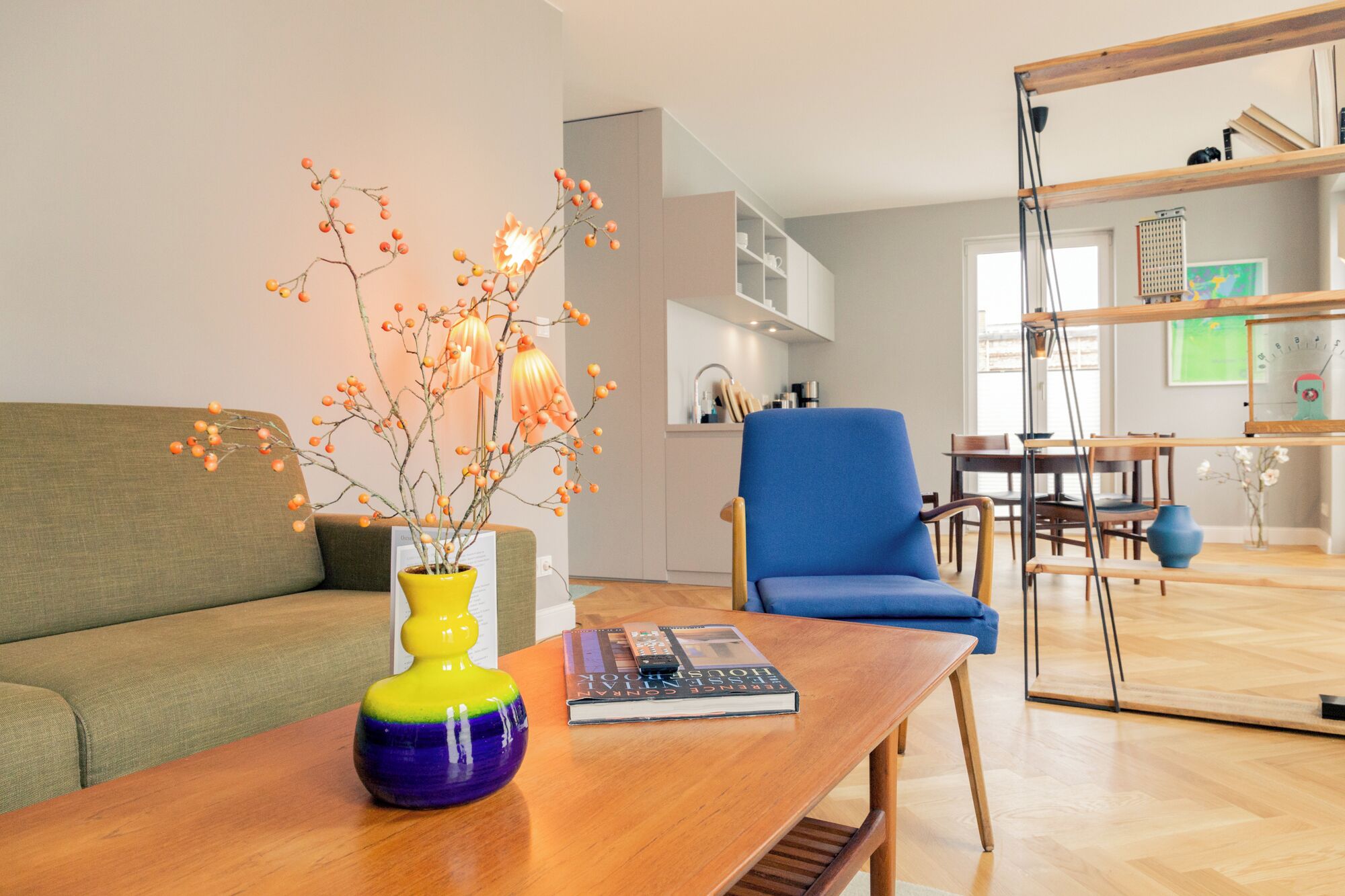 Ein modernes Wohnzimmer mit einer Couch, einem blauen Sessel, einem Holztisch, einer Vase mit Zweigen und einem Buch.