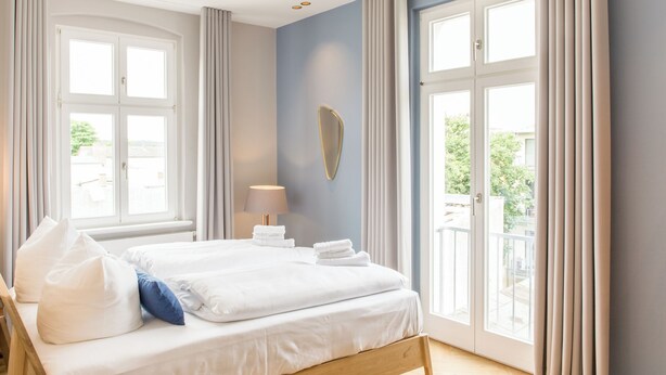 A bedroom with a wooden bed, white bedding, a bedside lamp, and large windows with curtains. - OSTKÜSTE - Nadler Hof Design Apartments (Heringsdorf)