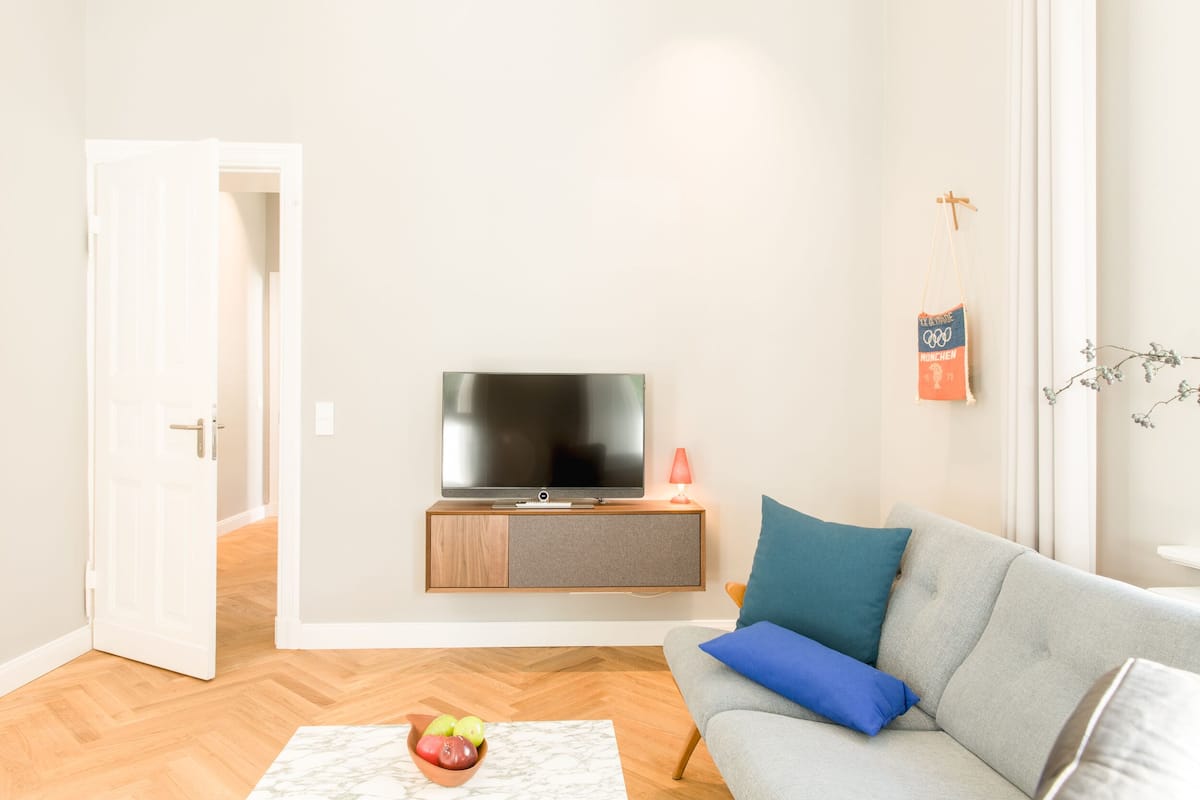 Un salon avec un canapé gris, un téléviseur posé sur un meuble en bois, un saladier de fruits sur une table et une porte donnant sur une autre pièce.