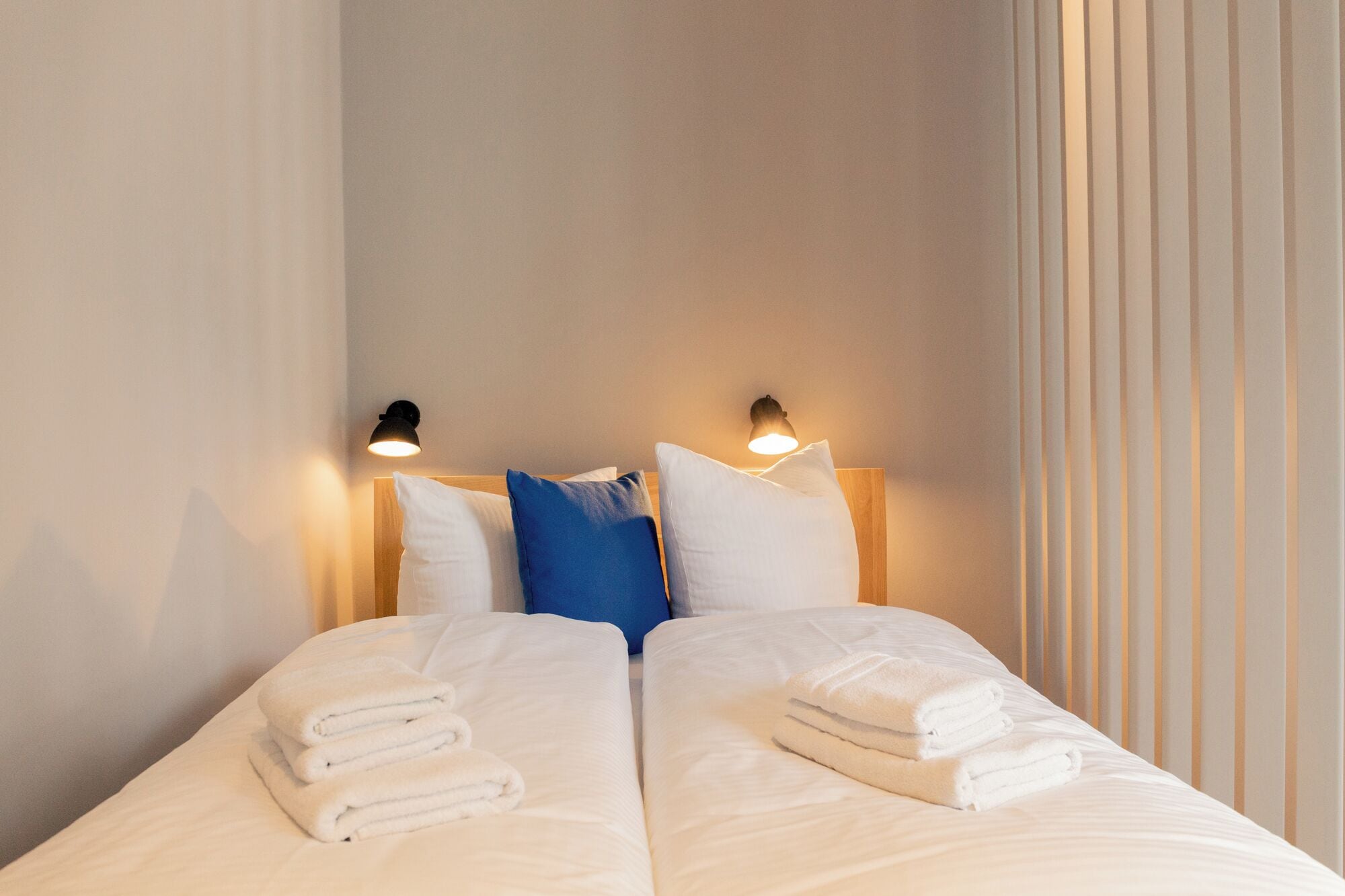 A neatly made double bed with white linens and folded towels, two bedside lamps, and a window with vertical blinds.