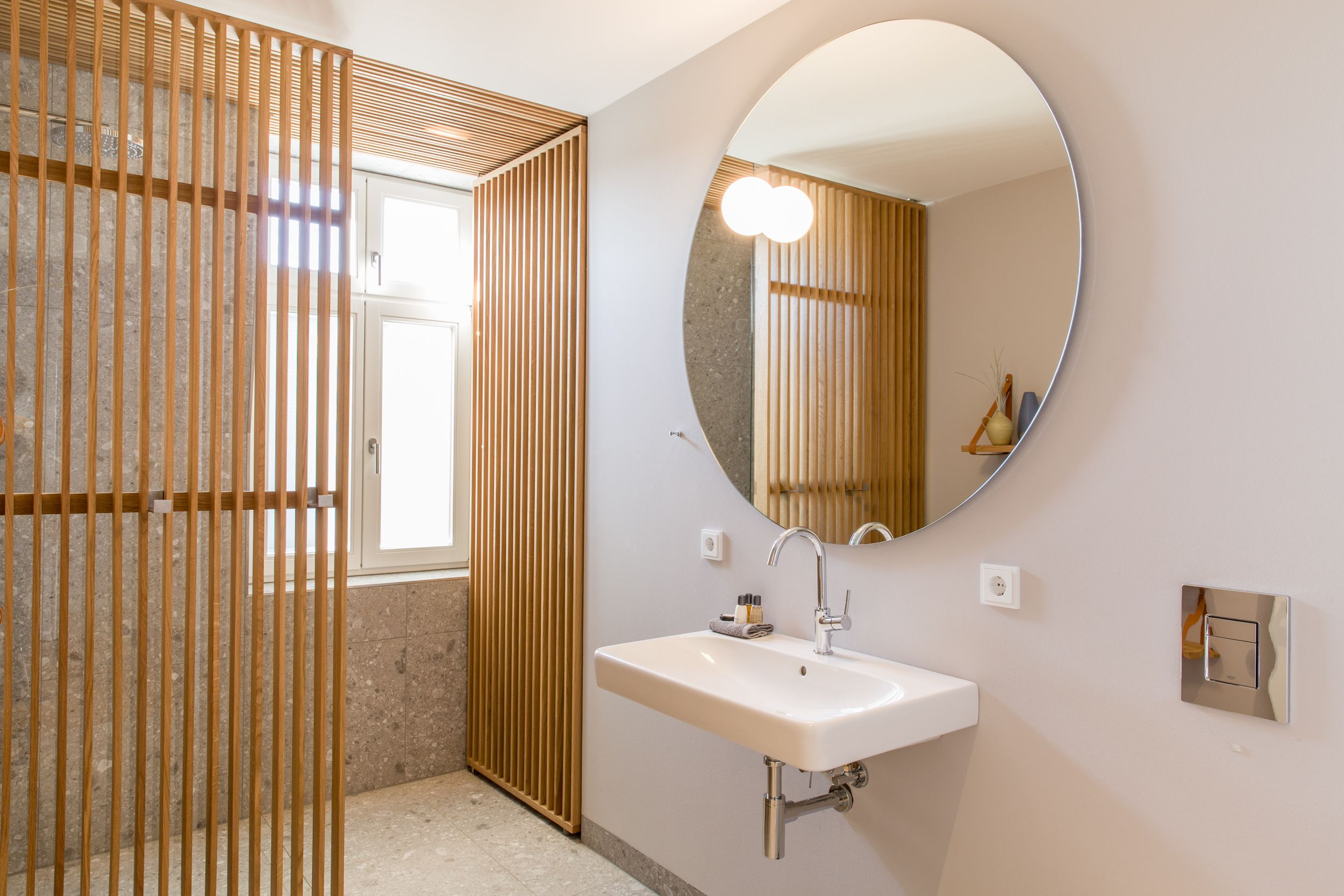 A bathroom with a white sink, a round mirror, and a wooden partition.