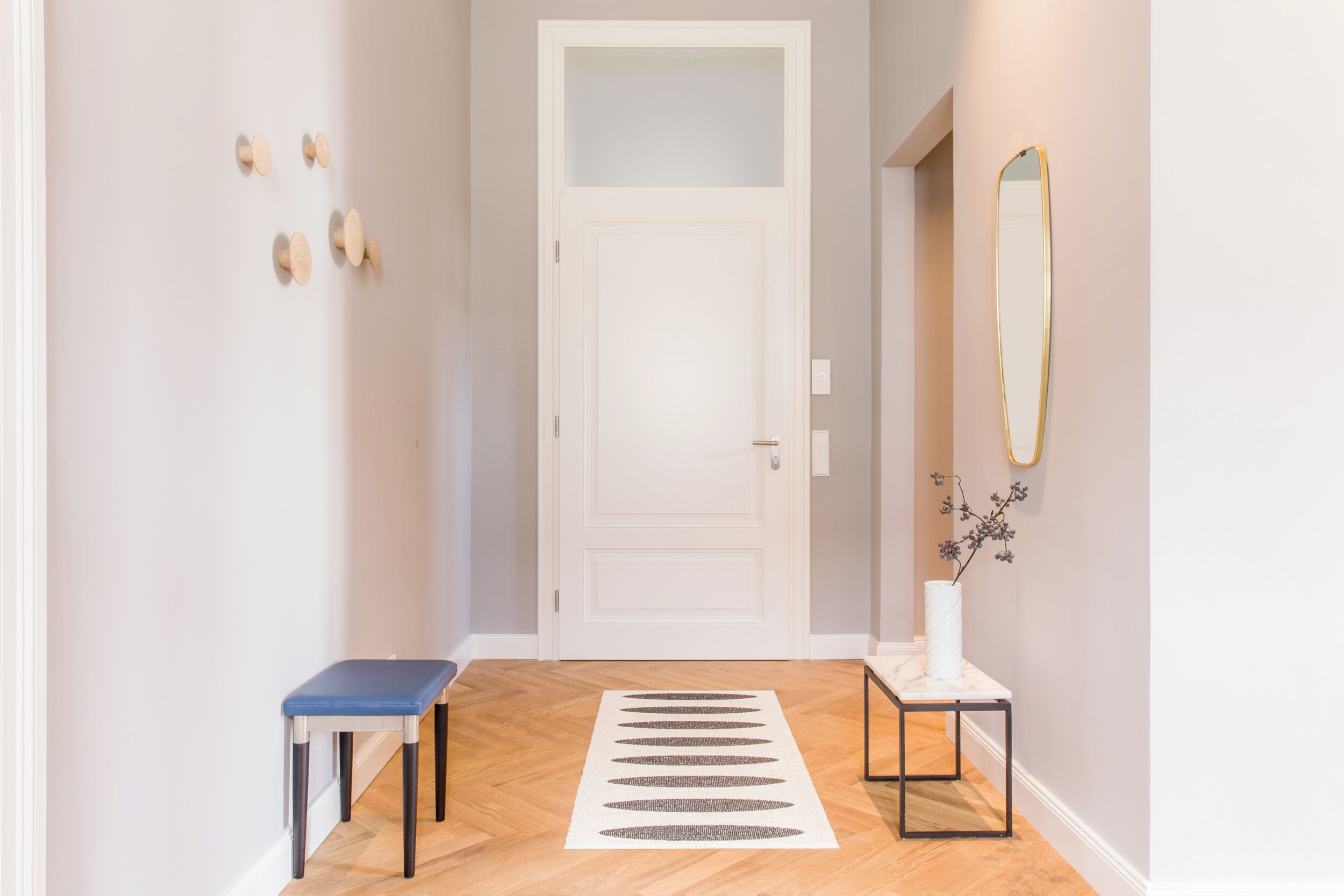 Un couloir avec une porte blanche, un banc bleu, une petite table avec un vase et un miroir au mur.