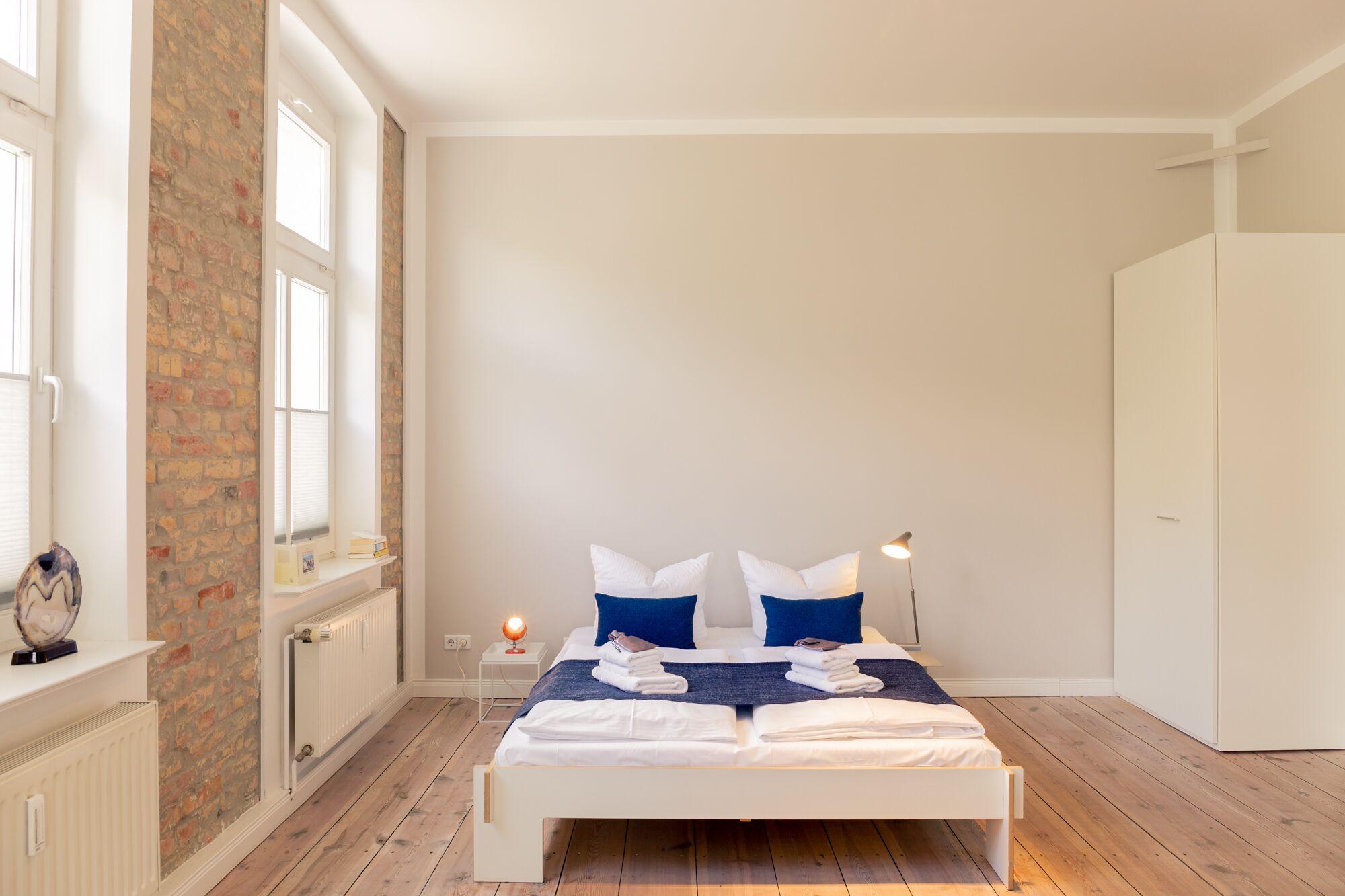 Un dormitorio con cama blanca, dos almohadas, una mesita de noche con lámpara, pared de ladrillo y una ventana.