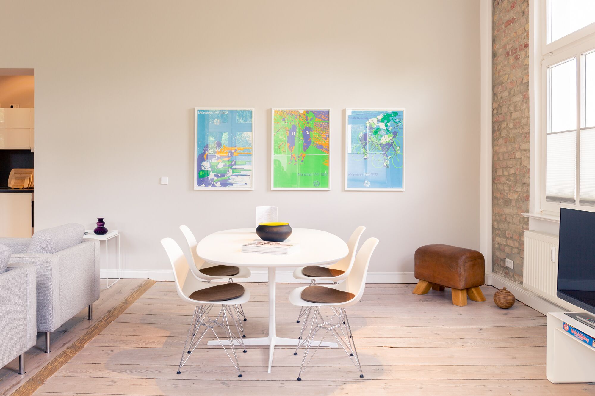 Un salon moderne avec une table à manger ronde, des chaises blanches, un canapé gris et trois tableaux abstraits aux couleurs vives accrochés au mur.