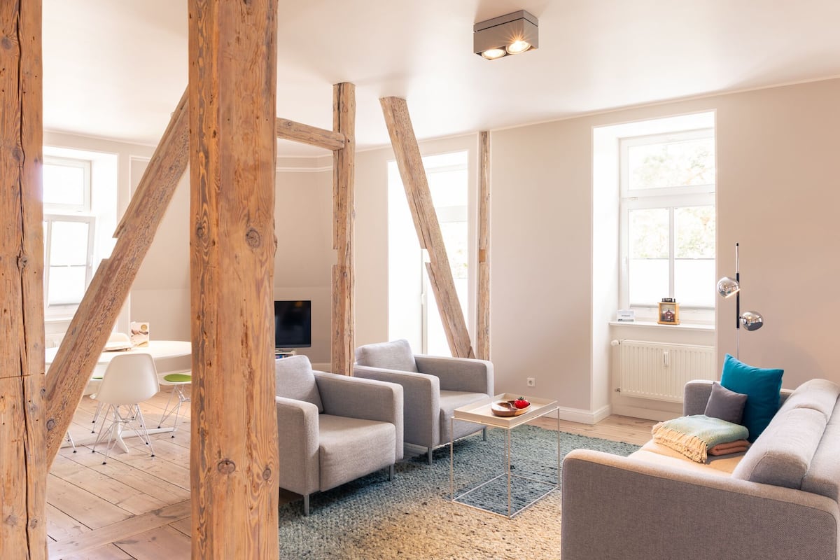 Een moderne woonkamer met zichtbare houten balken, een grijze bank, een klein tafeltje met een schaal, en een televisie op de achtergrond.