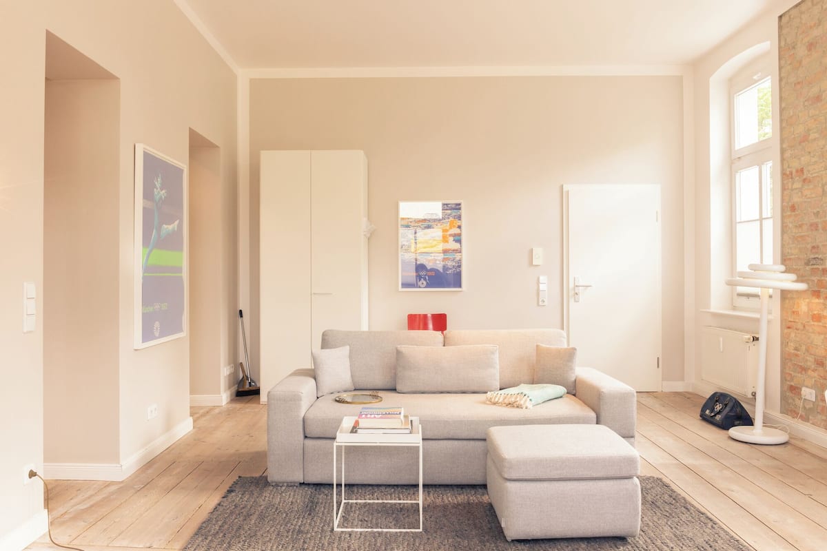Een moderne woonkamer met een grijze bank, een witte salontafel en een bijpassend voetenbankje. Er hangt een lijst met een foto aan de muur, er staat een rode fauteuil en een vloerlamp.
