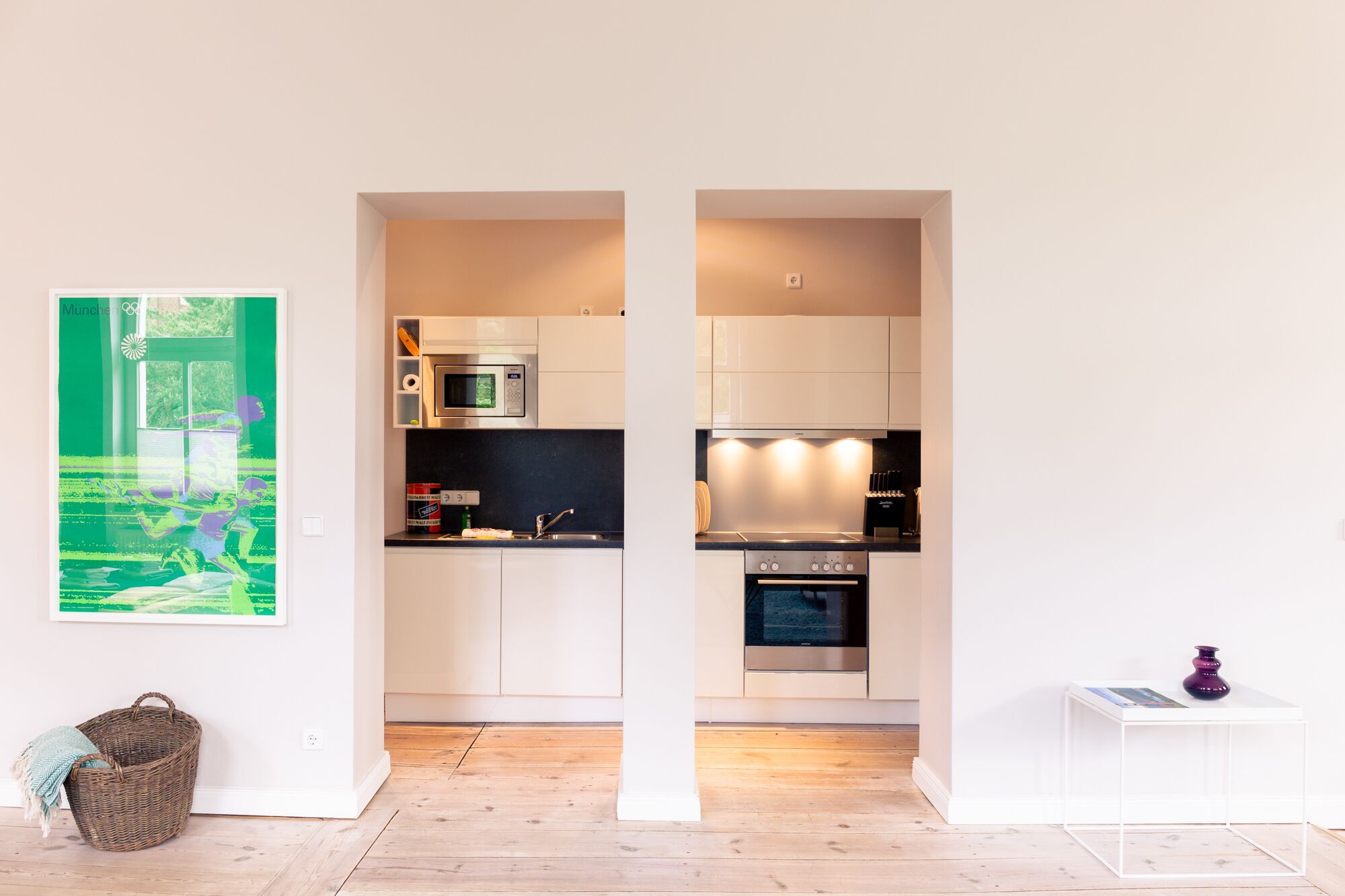 Una cocina moderna con armarios blancos, un microondas, una estufa y un fregadero. Hay un cuadro en la pared y una cesta en el suelo.