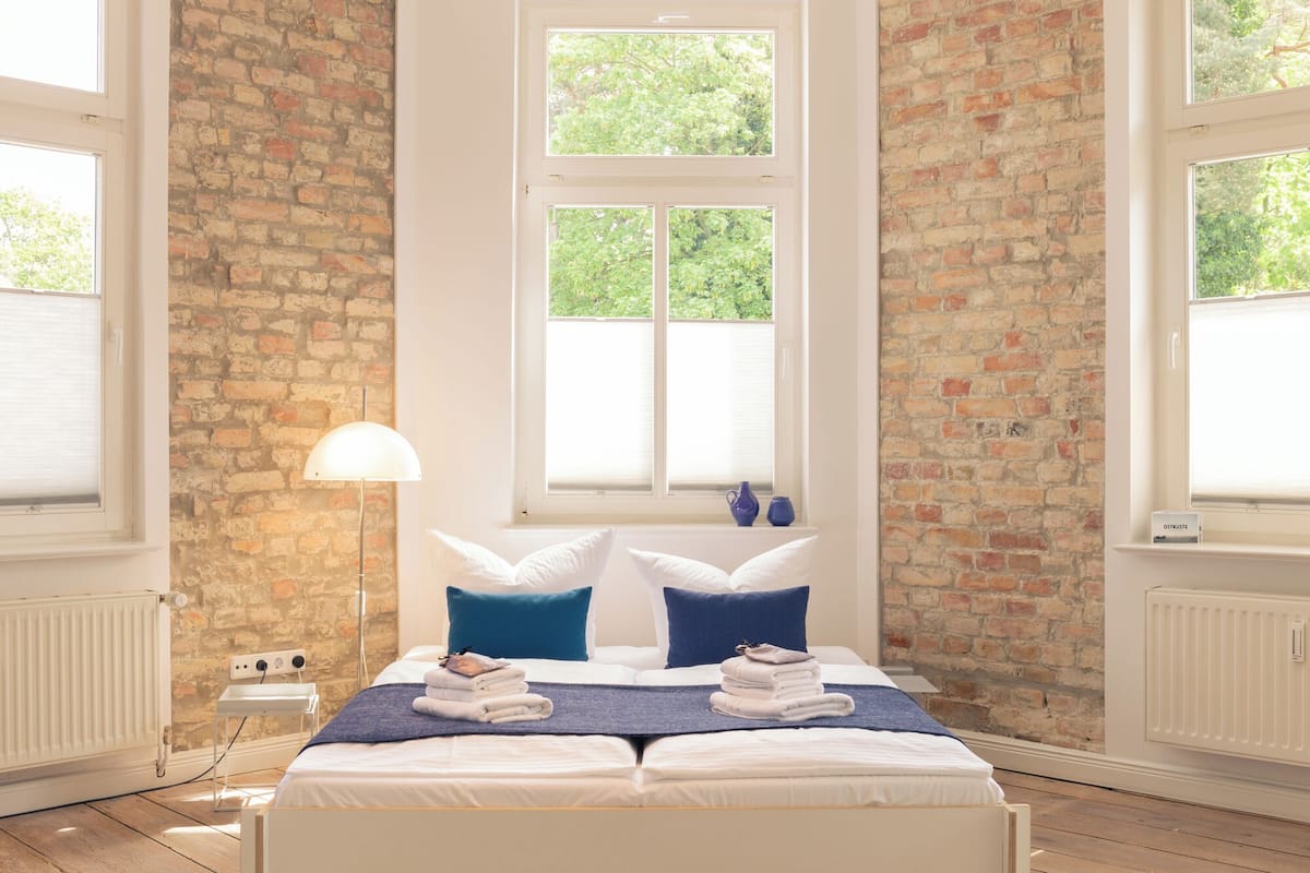 Een slaapkamer met een bakstenen muur, een groot raam, een bed met witte beddengoed, blauwe kussens en een lamp.